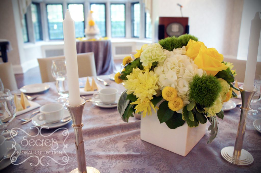 Low-profile centrepiece with fresh yellow roses, yellow cushion mums, green trick dianthus, light green carnations, succulents, cream hydrangea, yellow button mums, and dusty miller, in white cube pot (front angle)