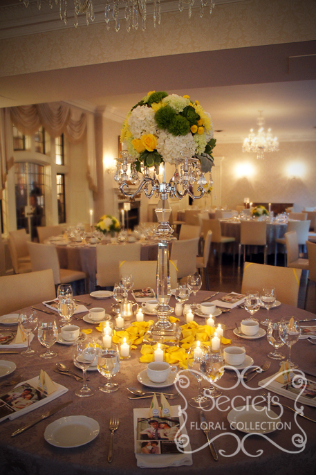 Head table centrerpiece of fresh yellow roses, yellow cushion mums, green trick dianthus, light green carnations, succulents, cream hydrangea, yellow button mums, and dusty miller, on real crystal candelabra. Surrounded with fresh yellow petals and votive candles