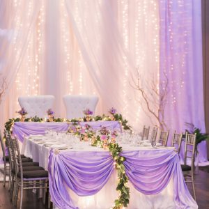 Wedding reception, with twinkle lights backdrop, and lavender head table. Dressed with purple floral and greenery garland. Toronto wedding flowers and decor at Fontana Primavera by Secrets Floral.